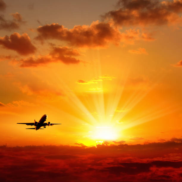 silhouetted flying commercial airplane over orange sunset sky with rays of sun