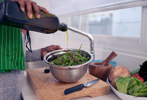 Close up image of female's hands pouring olive oil over green leaves salad in a bowl. The Woman adding olive oil into a fresh salad, preparing food on the kitchen countertop.
