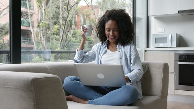 Black woman enjoying a hot chocolate while browsing on social media sitting down relaxed on couch at home