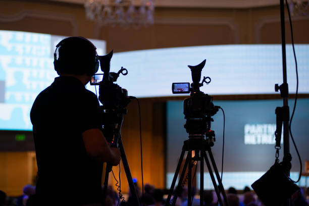 Camera Man Capturing Onstage Event A camera man with two cameras frames his shot for the presentation about to take place on the stage. He is somewhat silhouetted and is wearing headphones. reportage stock pictures, royalty-free photos & images