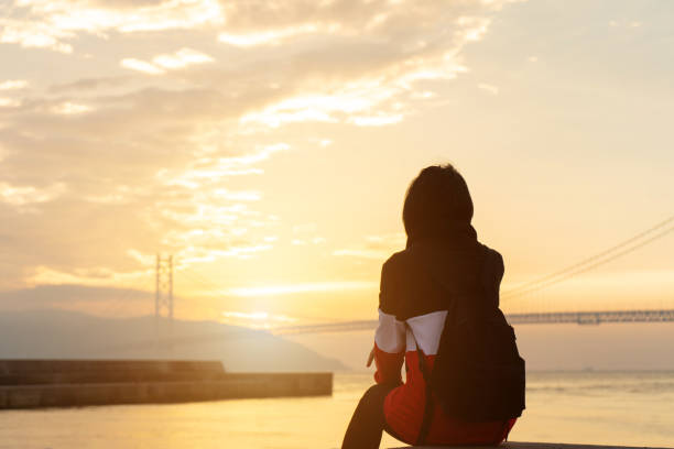 istock Silhouette alone young woman relax siting on riverside ocean and watching sunset and cloud sky with akashi kaikyo bridge. Thinking and concentrate in summer background with copy space for label text. 1211663869