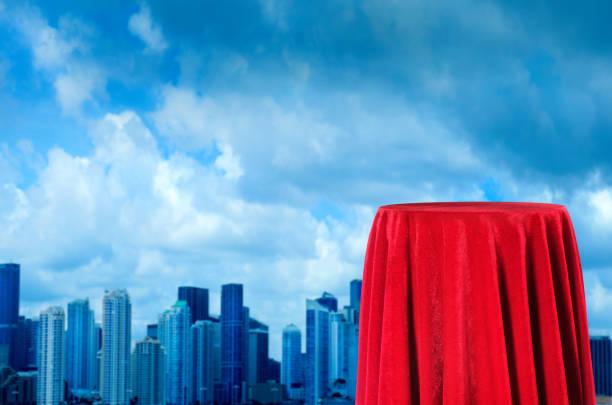 Conceptual close up empty red velvet covered pedestal with copy space for displaying products over cityscape of tall buildings with cloudy sky