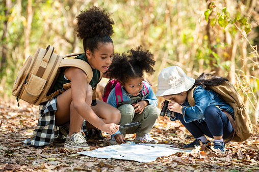 Group of happy pretty little girls hiking together with backpacks and sitting on forest dirt road with looking at the map for exploring the forest. Three kids having fun adventuring in sunny summer day