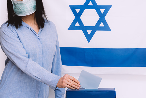 Israeli woman with face medical protected mask putting ballot in a ballot box on election day on Israel flag background.Israelis quarantined for coronavirus will get voting booths for March election.