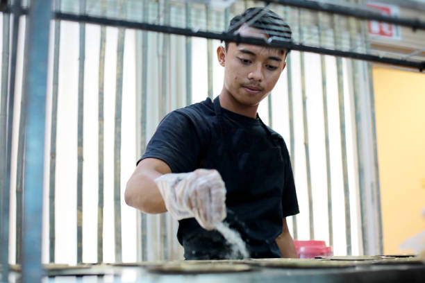 South East Asia: Small Business - Malaysian Local Food 'Apam Balik' A focus scene on human hand spreading sugar on 'apam balik' (a Malaysian favourite half flipover thin and crisp pancake with corn, sugar and Nestum - a Malaysian loved cereals) on multiple of mini flat pans. small business malaysia stock pictures, royalty-free photos & images