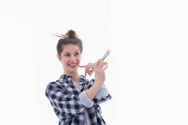 A beautiful young woman sporting paintbrushes on her hair bun holding a paintbrush on her hand in an assessing gesture on a light background. Artistic concept.