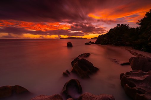 colorful sunset sky over tropical island beach with some rocks in the calm ocean on peaceful evening without people, deserted sand beach, long exposure