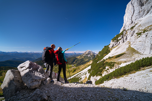 rear view mountaineer couple with backpacks standing near rocks high up in european mountains alps, woman pointing with hiking pole at rockface on sunny day with clear blue sky