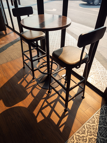 Cafe decoration design. Round wooden table with two modern bar stools on wooden floor near the glass window near brick wall on sunshine day vertical style.