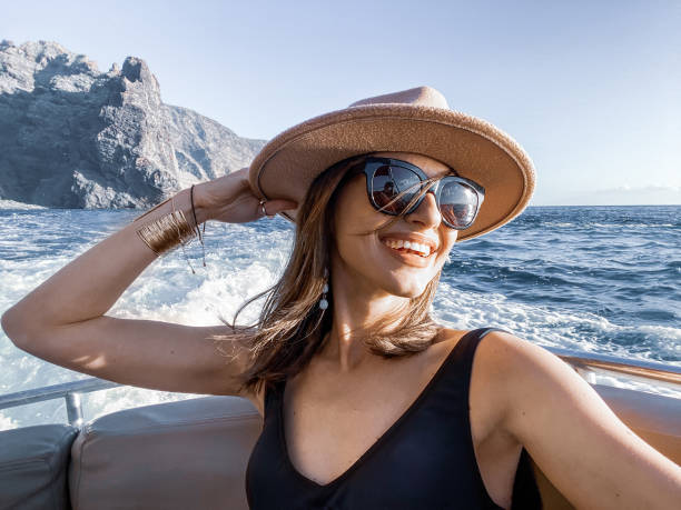 Woman enjoying ocean voyage Woman in swimsuit and sun hat enjoying ocean voyage, making selfie photo on phone with beautiful rocky coast on the background yacht selfie stock pictures, royalty-free photos & images