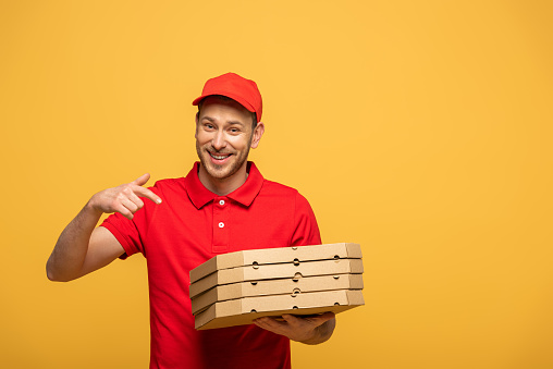 happy delivery man in red uniform pointing with finger at pizza boxes isolated on yellow