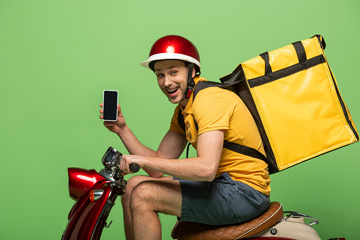 side view of happy delivery man in yellow uniform with backpack showing smartphone with blank screen on scooter isolated on green