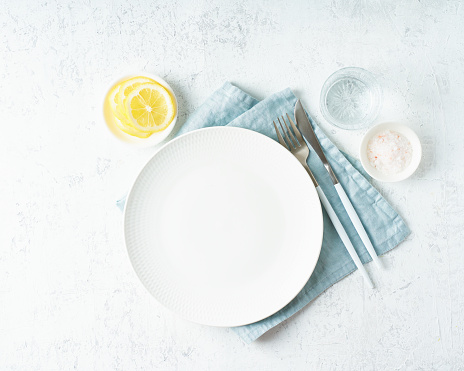 Clean empty white plate, glass of water, fork and knife on white stone table, copy space, mock up, top view. Concept for menu with utensil