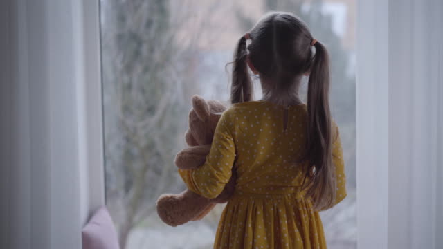 Back view of little Caucasian girl hugging teddy bear and looking out the window. Brunette kid spending day at home with toy. Leisure, lifestyle, childhood.