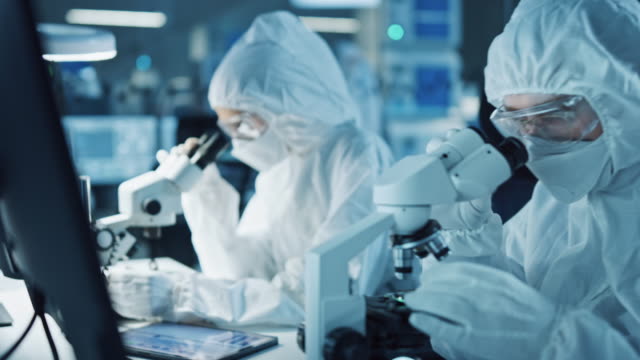 Research Factory Cleanroom: Engineers / Scientists wearing Coveralls and Gloves Use Microscopes to Inspect Motherboard Microprocessor Components, Developing High Tech Modern Electronics