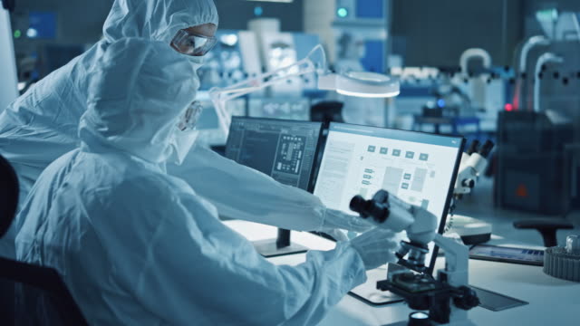 Research Factory Cleanroom: Engineer and Scientist Wearing Coveralls Talk and Work on Computer, Use Microscope to Inspect Motherboard Microprocessor, Develop Electronics for Medical Electronics