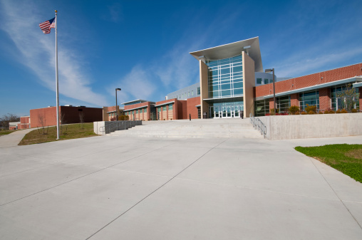 this color image is of a School Building or Business Building with American Flag. the building could also be a library or mall or church or university or gymnasium. the architecture is very modern. there is a massive sidewalk or walkway to the building. and the entrance is made of glass. the building is made of brick, glass and cement. there is also a lawn that is of green grass. and the lighting is natural sunlight. and the background is blue sky with white clouds. 