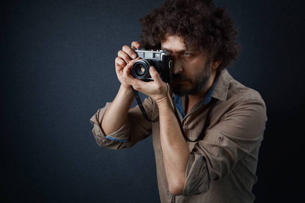Bearded man with curly hair holding old-fashioned camera and looking through viewfinder while standing against dark gray background