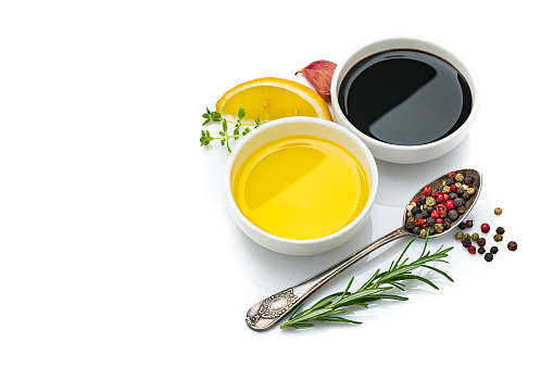 Cooking and seasoning: extra virgin olive oil and balsamic vinegar in white containers isolated on reflective white background. Rosemary twigs, garlic cloves, a lemon slice and mixed peppercorns are around the containers and complete the composition. The composition is at the right of an horizontal frame leaving useful copy space for text and/or logo at the left. Predominant colors are yellow, black and white. High resolution 42Mp studio digital capture taken with Sony A7rII and Sony FE 90mm f2.8 macro G OSS lens