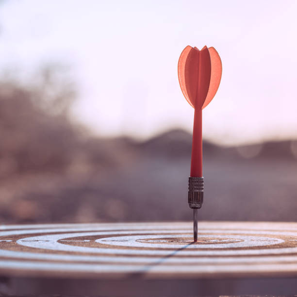 Close up red dart arrow hitting target centre dartboard on sunset background. Business targeting and focus concept.