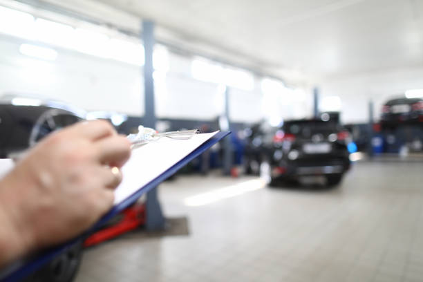 Close-up of worker filling information about damaged vehicle. Male holding pen and clipboard. Garage full of automobiles. Service station and car maintenance concept