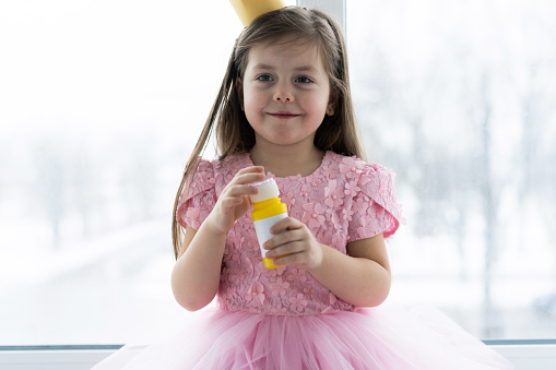 Little cute girl in beautiful dress is sitting near the window at home and blowing soap bubbles