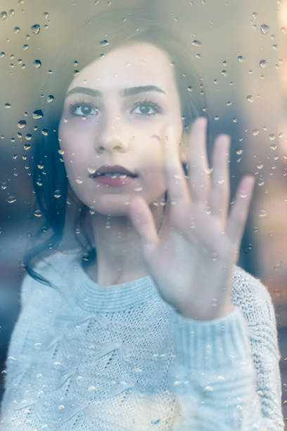 istock Young Woman Looking Through Window With Raindrops 1211807145