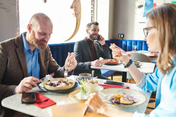 Business people in cafe: successful young businessman eating salad and discussing business on phone while colleagues having lunch together