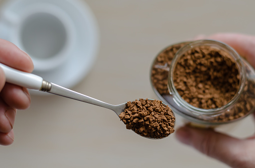 Glass jar with coffee in a hand. A hand holds a spoon full of instant coffee. Pile of instant coffee grains in metal spoon over glass jar. Caffeine drink.  View from above. Selective focus. Close-up.