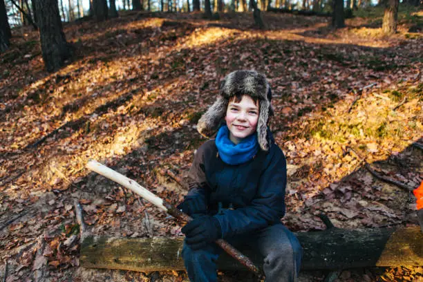December 1, 2018 - Kampinos, Poland: cheerful beautiful pre-adolescent boy sits near dirt road in a pine forest in the evening with sun behind the trees, looking at camera, smiling, shaving his stick with a knife, being healthy, happy and active