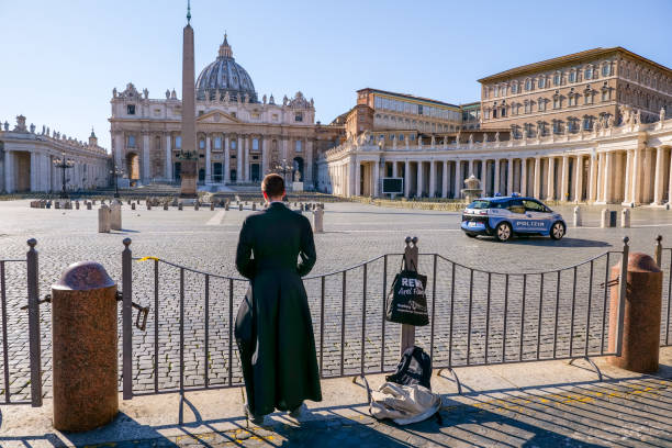 A priest prays in St. Peter's Square closed and deserted due to the Covid-19 lockdown Vatican, Italy, March 11 -- A Catholic priest prays in front of the square of St. Peter's Basilica, empty and closed to visitors and tourists for the coronavirus emergency in Italy and controlled by the police. church of san pietro photos stock pictures, royalty-free photos & images