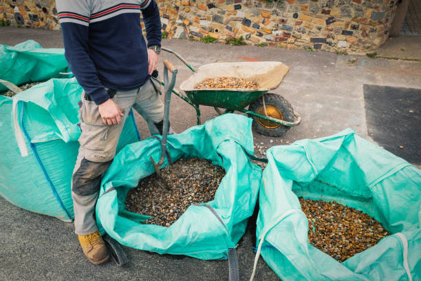 Large open sacks of gravel pebbles for a garden design project delivered to the kerb side. A man in gardening cloths stands by them near the shovel and wheelbarrow used to move them to the house. grandes sacos abiertos de guijarros de grava para un proyecto de diseño de jardín entregado al lado del bordillo. un hombre con paños de jardinería está junto a ellos cerca de la pala y la carretilla utilizada para trasladarlos a la casa.