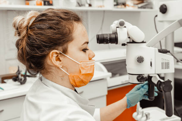 giovane donna dentista medico guarda attraverso un microscopio professionale in una clinica dentale. un medico con una maschera medica usa e getta e un berretto. attrezzature avanzate in odontoiatria - 5934 foto e immagini stock