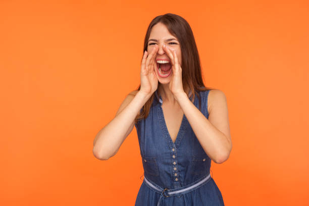 femme de brunette dans la robe de denim gardant des bras autour de la grande bouche ouverte, criant fort - mouth open women displeased anger photos et images de collection