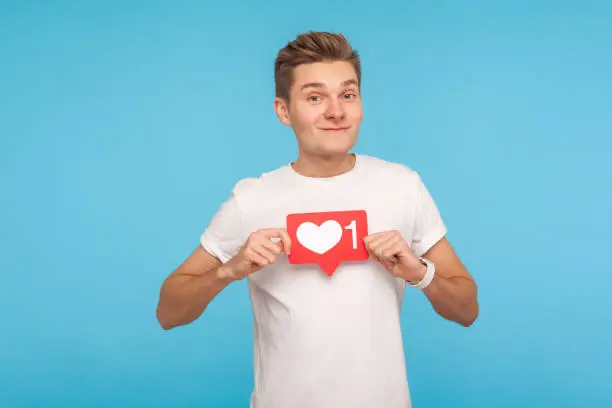 Photo of Positive smiling man in casual white t-shirt holding social media button, showing heart like icon