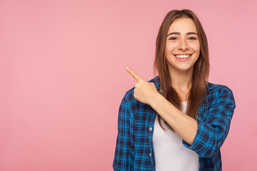 Look at advertisement! Portrait of charming brunette girl in checkered shirt pointing aside at empty place for ad content. studio shot isolated on pink background, copy space for commercial product