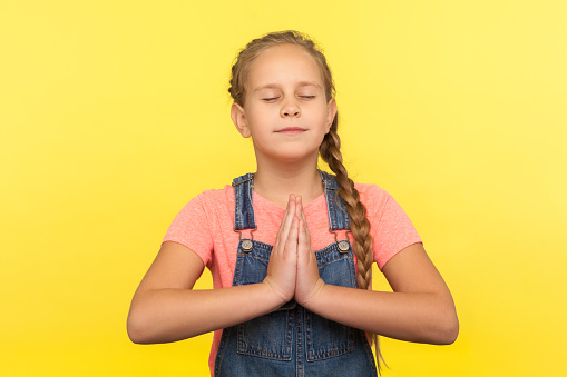 Yoga and inner harmony. Portrait of cute little girl with braid in denim overalls praying with closed eyes, feeling calm relaxed with peaceful mind. indoor studio shot isolated on yellow background
