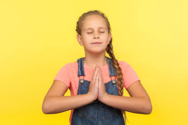 yoga and inner harmony. portrait of cute little girl with braid in denim overalls praying with closed eyes - praying girl imagens e fotografias de stock