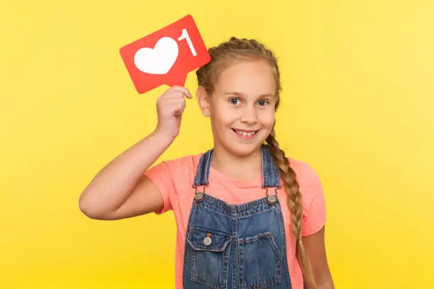 Photo of Like and comment for child blogging. Portrait of happy little girl in denim overalls holding social network heart button