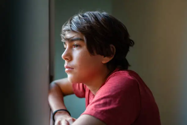Photo of latinx preadolescent boy looking out through window, reflecting, relaxing, pensive.