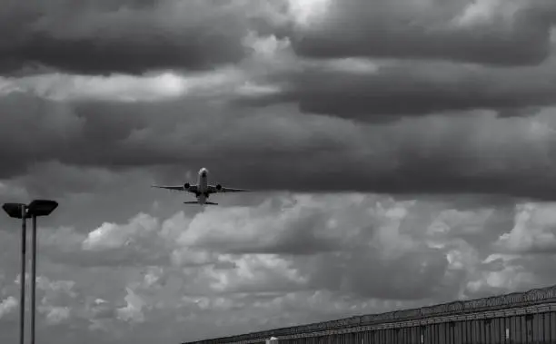 Photo of Black and white scene of airplane landing at the airport. Commercial airline flying on dark sky and white clouds over fence and lamp post. Aviation business crisis concept. Journey vacation flight.