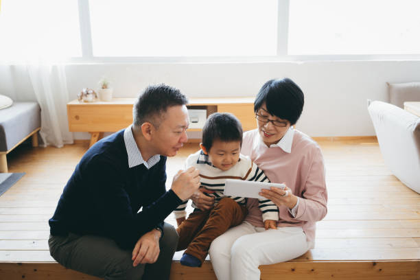 fröhliche asiatische großeltern sitzen auf dem boden im wohnzimmer mit digitalen tablet mit kleinen enkel zusammen zu hause - senior adult digital tablet domestic life learning stock-fotos und bilder