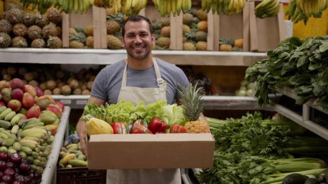 Friendly man preparing a delivery for customer in a cardboard box full of fresh fruits and vegetables looking at camera smiling
