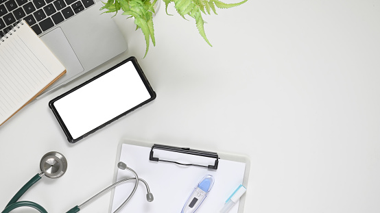 Top view image of workspace. Flat lay white blank screen smartphone, clipboard, digital thermometer, stethoscope and potted plant on white working table. Orderly doctor workplace concept.