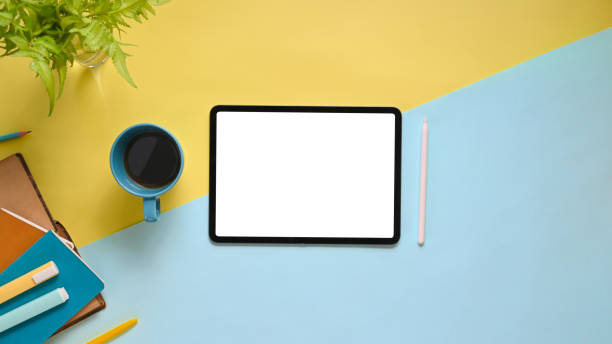 Top view image of workspace with flat lay white blank screen computer tablet, coffee cup, stylus pen, marker pens, stack of notebook and potted plant on colorful working table.