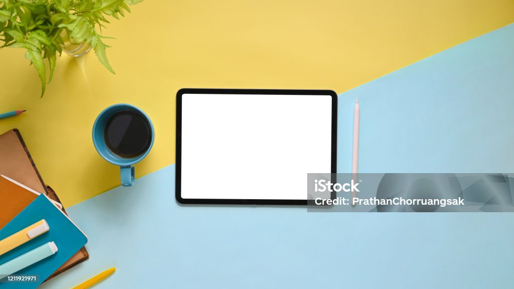 Top view image of workspace with flat lay white blank screen computer tablet, coffee cup, stylus pen, marker pens, stack of notebook and potted plant on colorful working table. Digital Tablet Stock Photo