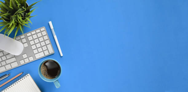 istock Top view Simple panorama workspace. Flat lay coffee cup, pens, pencil, notebook, keyboard, wireless mouse and potted plant on modern blue working table. Orderly workplace concept. 1211921991