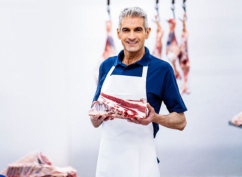 Portrait of smiling mature man working in slaughterhouse. Male butcher is holding fresh raw meat. He is at food processing plant.