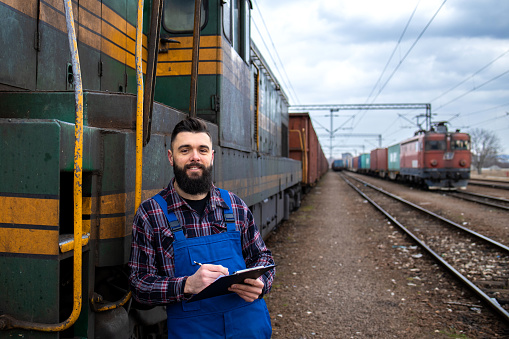 Portrait of engine train driver standing by locomotive at train station and holding departure schedule. In background railroad and cargo containers.