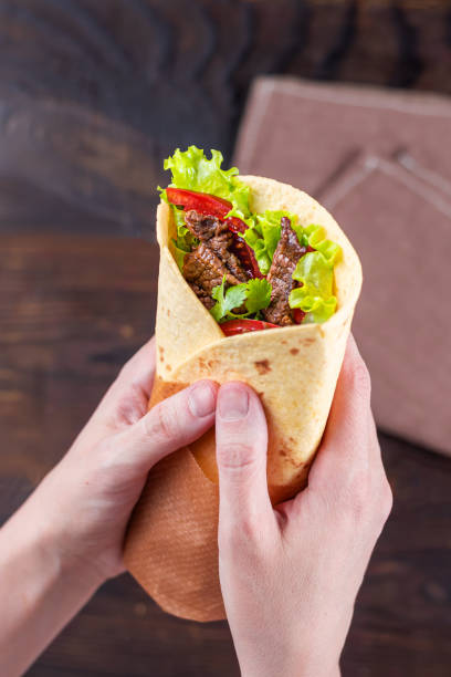 Burrito with beef in female hands Burrito with slices of fried beef, tomato, lettuce, wrapped in cheese tortilla in female hands. Mexican national cuisine. Food on the background of a wooden table. Vertical view. burrito photos stock pictures, royalty-free photos & images